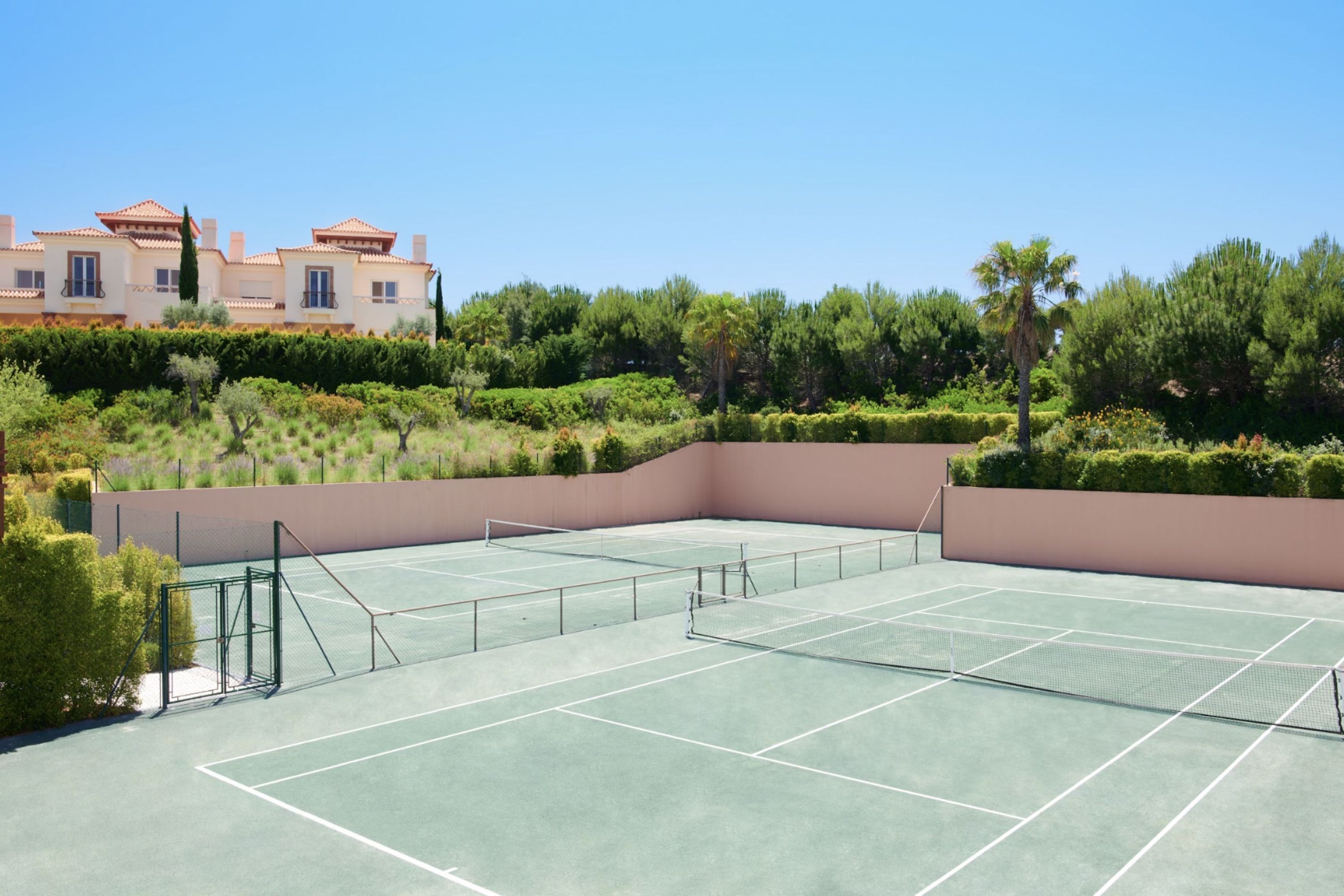 Una cancha de tenis con una red, rodeada de vegetación y un edificio al fondo.