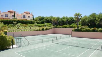 Una cancha de tenis con una red, rodeada de vegetación y un edificio al fondo.