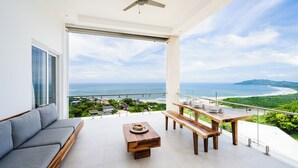 A balcony with a sofa, wooden table, and bench, overlooking a coastal view with a beach and greenery. - La Vista #601, brand New 3 bd Condo overlooking Tamarindo Bay (Playa Tamarindo)