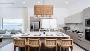 A modern kitchen with a central island, four wicker chairs, a hanging light fixture, and a view of the sea through large windows. - La Vista #601, brand New 3 bd Condo overlooking Tamarindo Bay (Playa Tamarindo)