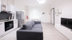 Cocina moderna con armarios blancos, horno integrado y refrigerador de acero inoxidable. En la sala de estar, un sofá gris, un televisor de pantalla plana sobre un pedestal blanco, y una mesa de comedor con sillas al fondo.