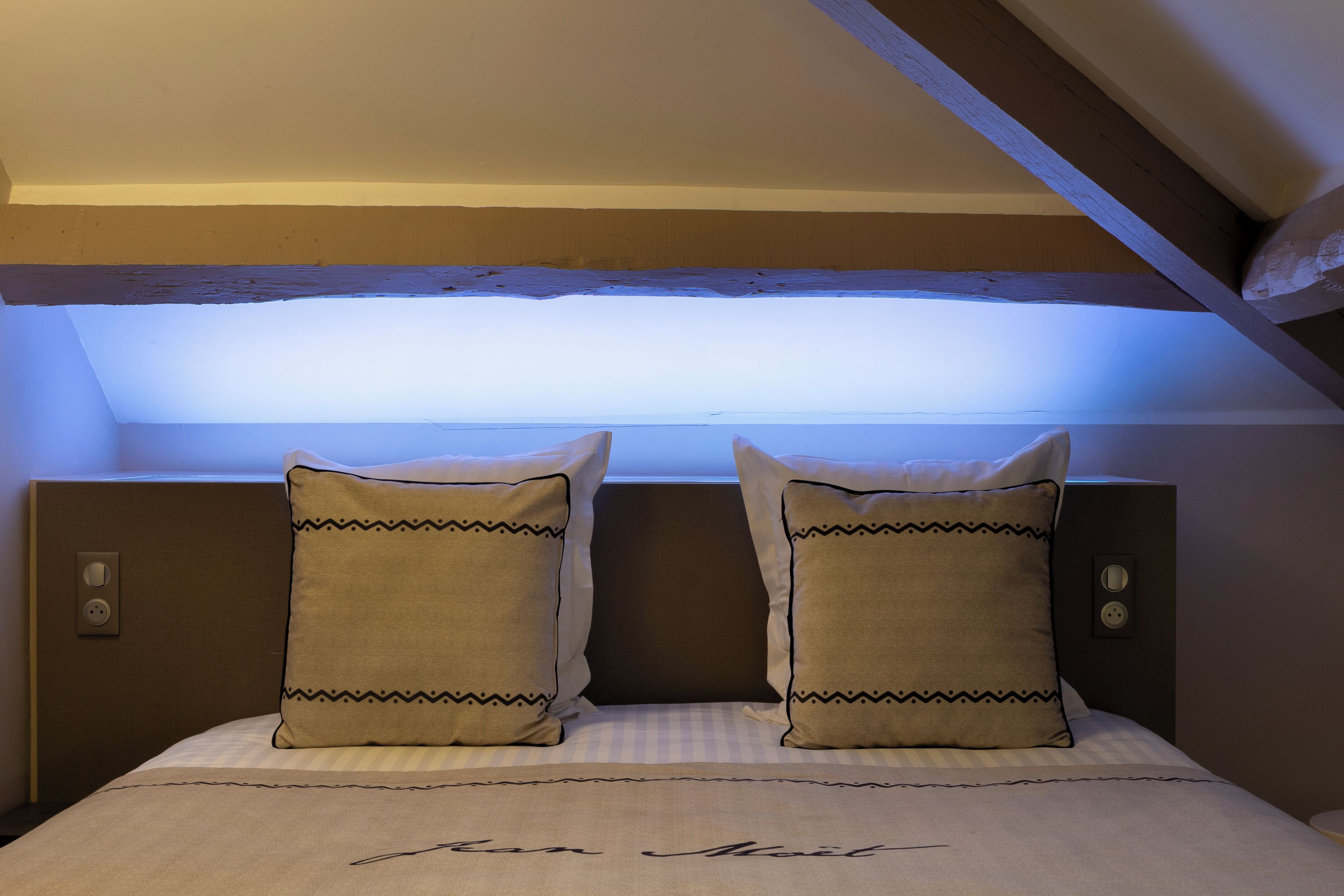 Quarto com teto inclinado, uma cama com duas almofadas e uma mesa de cabeceira com tomada.
