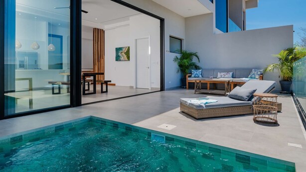 Ein moderner Poolbereich im Freien mit einer verglasten Terrasse, einem Essbereich mit Tisch und Stühlen sowie einer Lounge-Ecke mit Kissen und einem kleinen Tisch.
