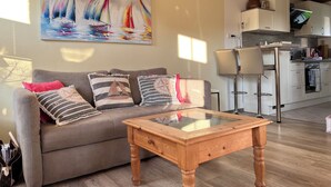 A living room with a sofa, cushions, a coffee table, and a painting of sailboats. - Reed-roofed FH with beach chair in the garden and close to the beach (Dranske)