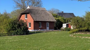 A red brick house with a thatched roof, surrounded by a green lawn and trees. - Reed-roofed FH with beach chair in the garden and close to the beach (Dranske)