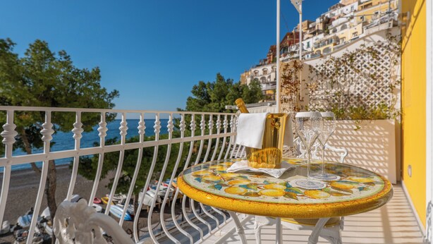 A round table with a lemon pattern, a bottle, and glasses on a balcony overlooking a beach and buildings. - La Sirenetta Luxury Suite Positano (Positano)