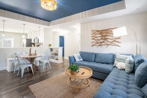 A modern living room with a sectional sofa, a dining area with white chairs, and a kitchen in the background. - Seashell Cove: Dogs Welcome! Ground Level - Vibrant Beach Cottage (Oak Island)