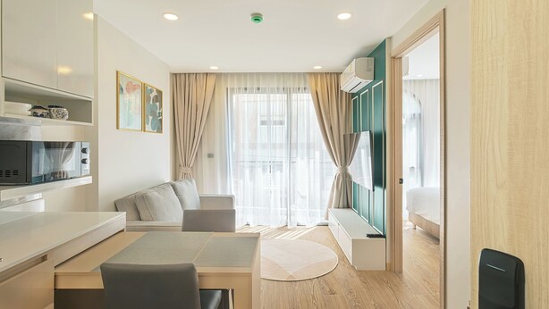 Una habitación de hotel moderna con kitchenette, zona de comedor, sofá y un amplio ventanal con cortinas.