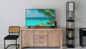 A wooden TV stand with a flat-screen TV displaying a beach scene, a black chair, and a black shelving unit with decorative items.