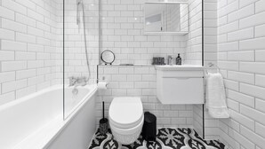 A modern bathroom with white subway tiles, a glass-enclosed shower, a white toilet, a black trash bin, and a patterned floor. - London Grove Apartments (London)