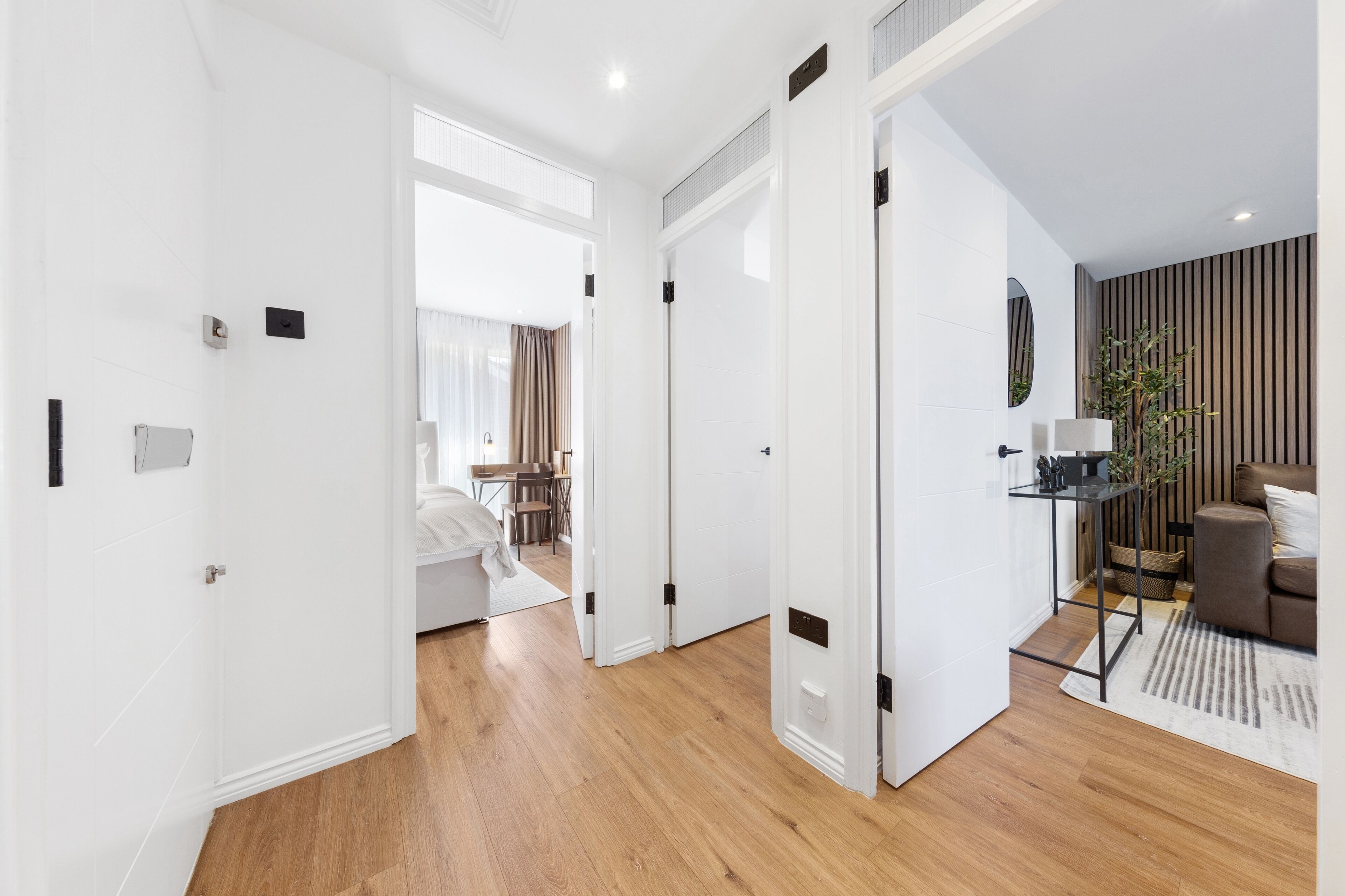 A modern hotel room with a wooden floor, white walls, and a dining area visible through an open door.