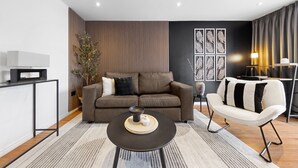 A modern living room with a brown sofa, a white armchair, a coffee table, and a side table with a lamp. - London Grove Apartments (London)