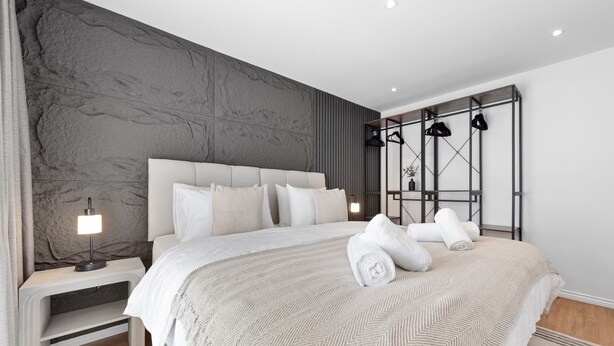 A modern bedroom with a large bed, a nightstand, a wall-mounted shelf, and a hanging light fixture. - London Grove Apartments (London)