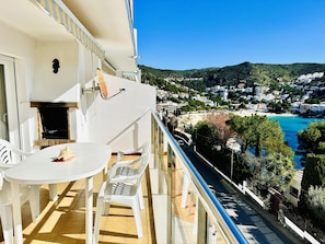Un balcon avec une table et des chaises blanches, donnant sur une ville côtière avec ses bâtiments et sa plage.