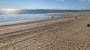 A wide sandy beach with footprints and tire tracks, a calm sea, and a clear sky.