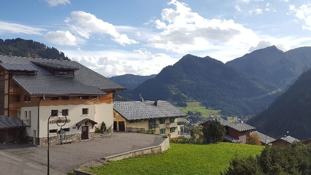 A mountain village with traditional buildings, a paved courtyard, and a view of the surrounding hills.