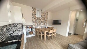 Eine moderne Küche mit gemusterten Fliesen, ein Essbereich mit Holztisch und -stühlen, ein an der Wand montierter Fernseher und ein graues Sofa im Wohnbereich.