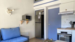 Eine kompakte Küchenzeile mit Mikrowelle, Kühlschrank und Herd. Ein blaues Sofa und ein Esstisch sind ebenfalls zu sehen.