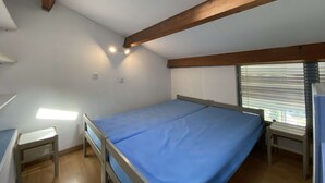 Ein kleines Zimmer mit einem Einzelbett, einem Dachfenster, Holzbalken, einem Fenster mit Jalousien und einem Nachttisch.