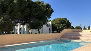 Une piscine rectangulaire, dotée de deux échelles en métal, est entourée d’un mur de couleur beige et de marches menant à un bâtiment agrémenté de balcons.