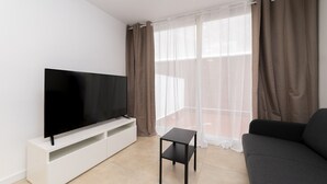 Een moderne woonkamer met een flatscreen televisie op een witte tv-meubel, een zwarte salontafel, een grijze bank en een groot raam met dunne gordijnen.