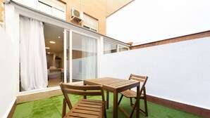 Een klein terras met een houten tafel en stoelen, een vloer van kunstgras en een witte muur.