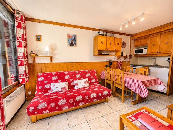 Una habitación acogedora con un sofá rojo estampado, armarios de madera, una pequeña cocina y una mesa de comedor con mantel a cuadros.