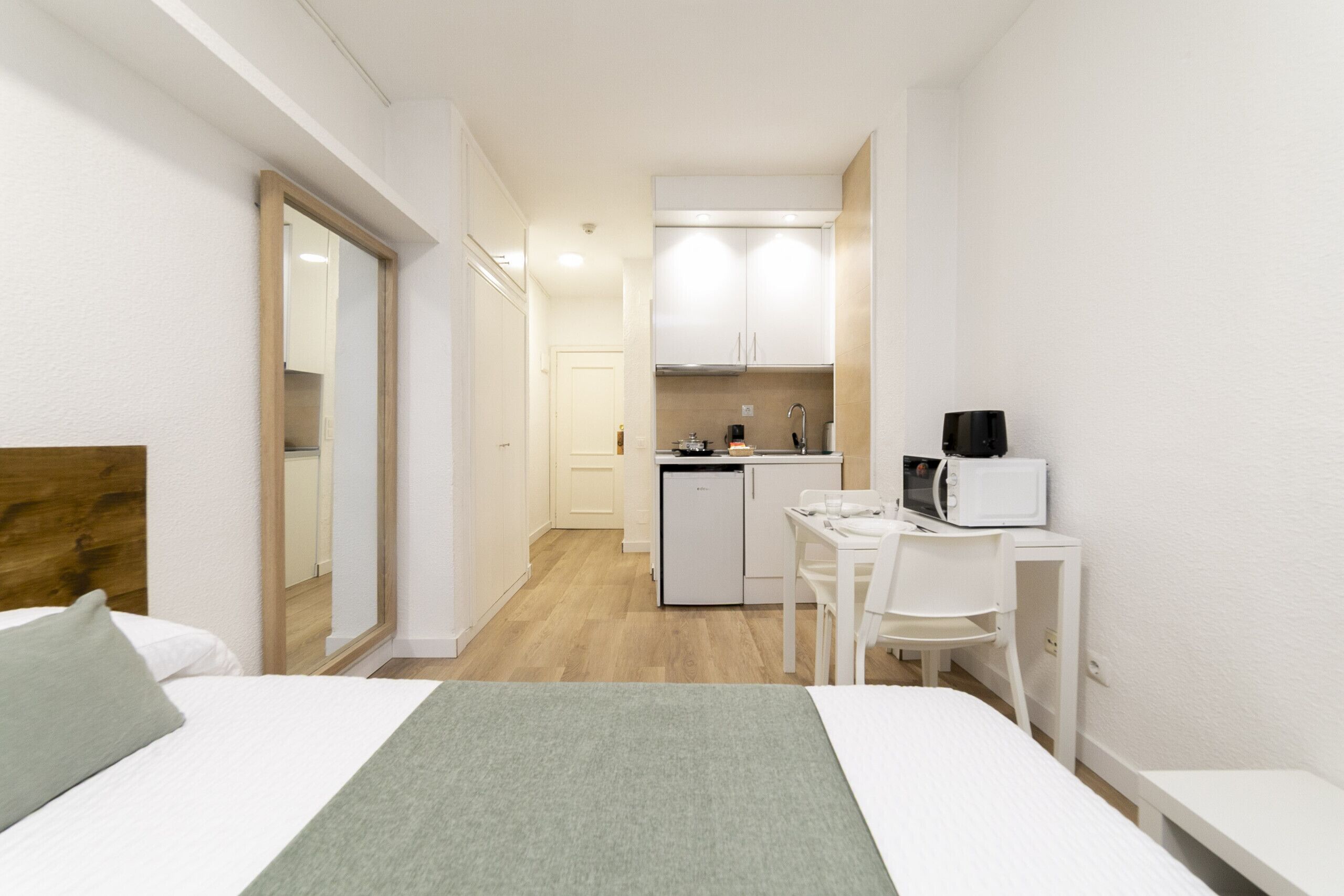 A compact hotel room with a bed, a small kitchenette, a dining area, and a mirror.