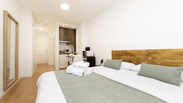Une chambre d’hôtel avec un grand lit, une tête de lit en bois, une kitchenette et une salle de bain visible à travers une porte ouverte.