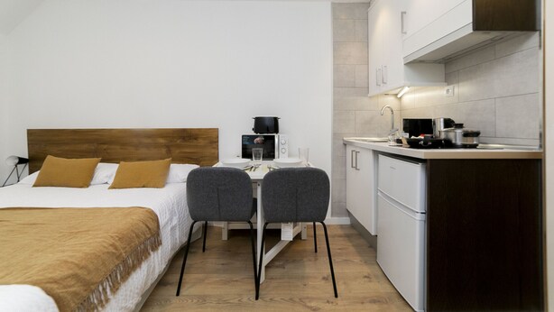 Une chambre d’hôtel de taille réduite comprenant un lit, une petite table à manger avec deux chaises, un coin cuisine et une tête de lit en bois.