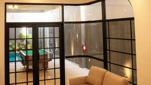 A modern living room with a sofa, a coffee table, and a wicker chair. There is a glass partition with a view of an outdoor pool.