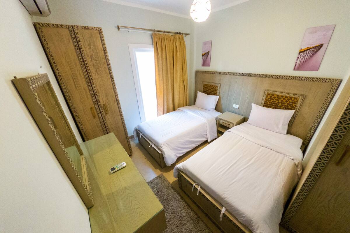 A hotel room with two beds, a bedside table with a remote control, a mirror, and a window with curtains.