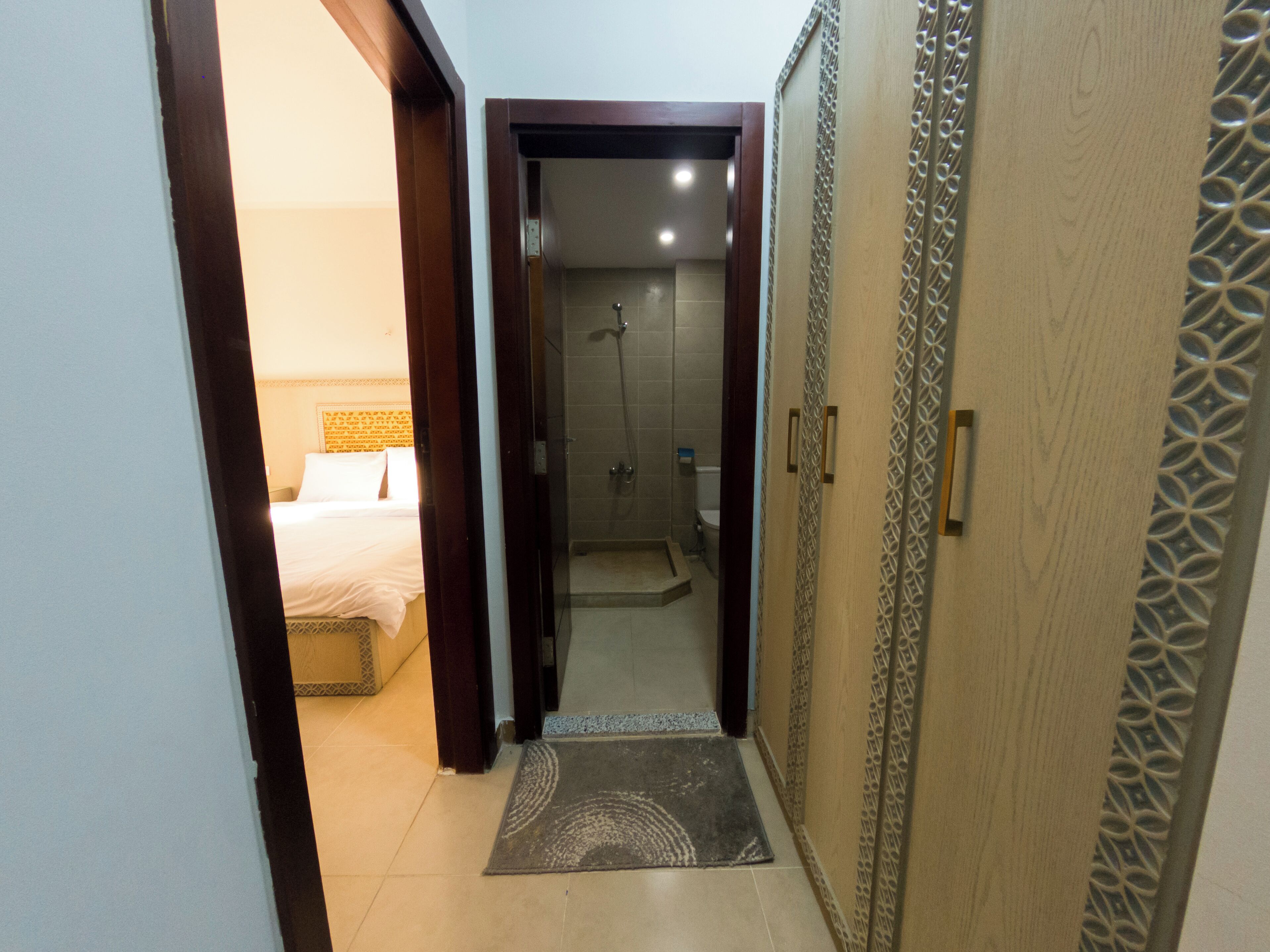 Pasillo de hotel que conduce a un baño con ducha y una cama con cabecera.