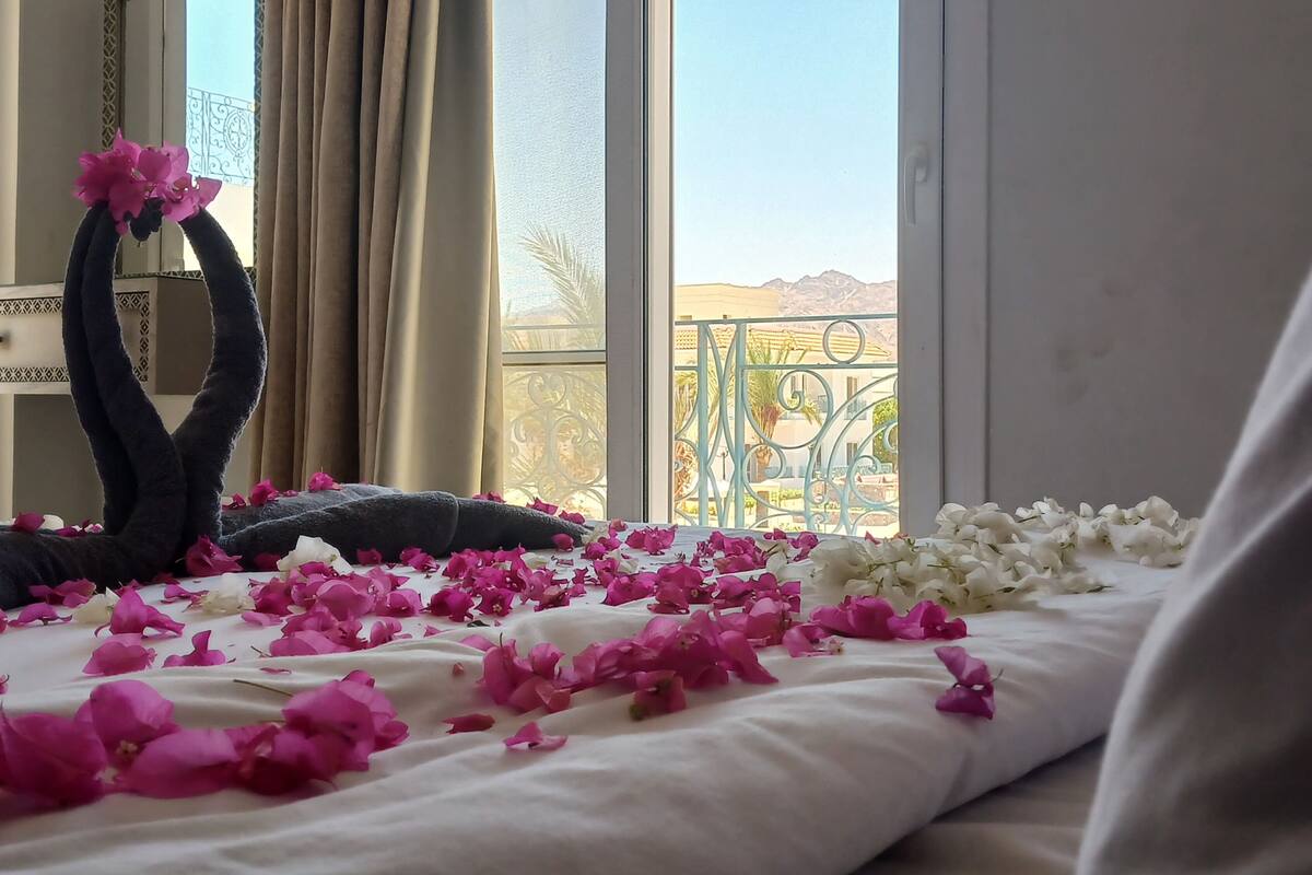 A hotel room with a bed adorned with pink and white flower petals, a decorative mirror, and a view of a balcony with a railing.