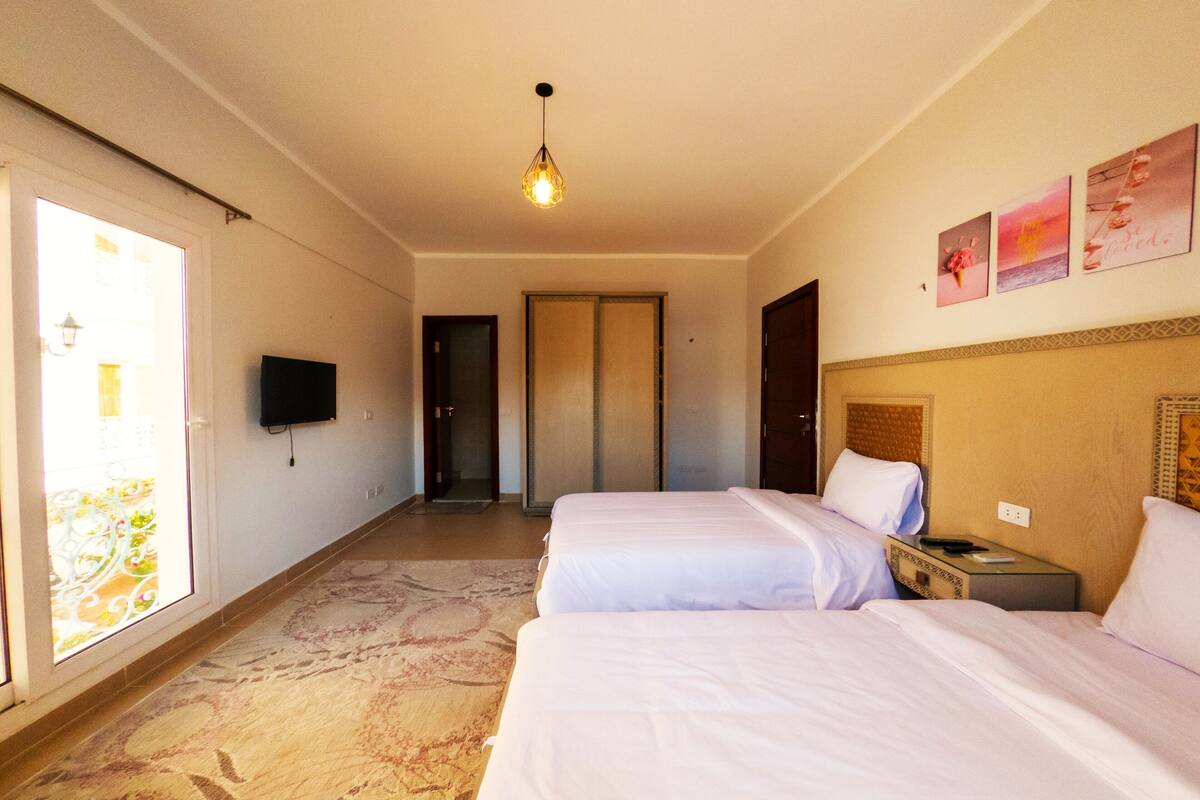 A hotel room with two beds, a television on the wall, and a door leading to a balcony.