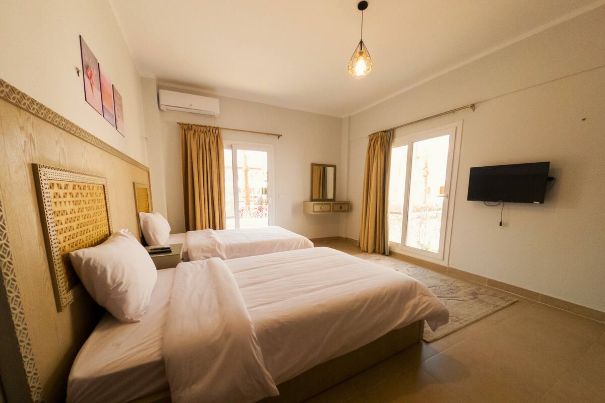 A hotel room with two beds, a flat-screen TV, a window with curtains, and an air conditioning unit.