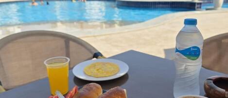A dining table with breakfast items including pancakes, fruit, and a coffee cup, overlooking a swimming pool with a view of a resort building.