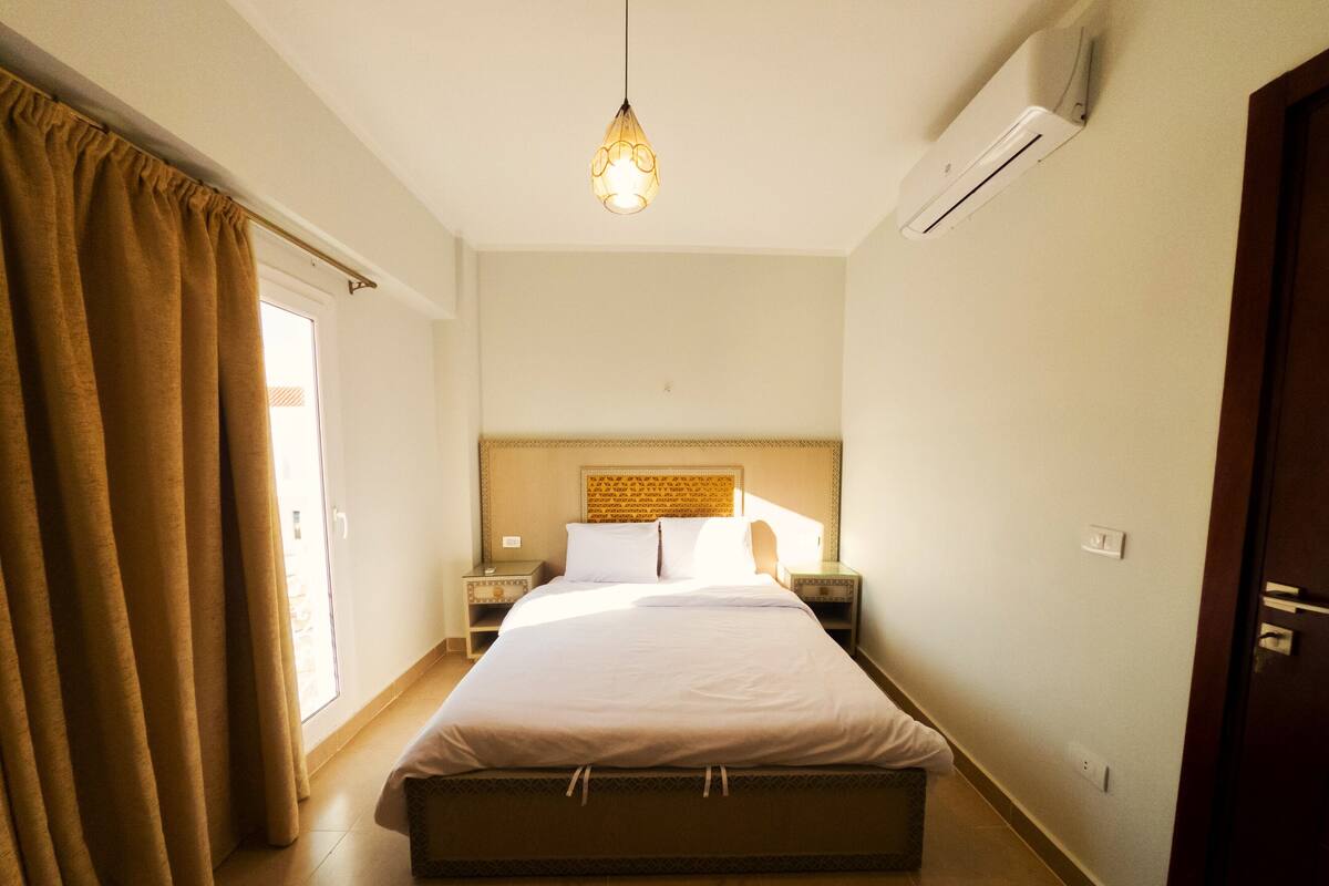 A hotel room with a bed, bedside tables, an air conditioner, and a hanging light fixture.