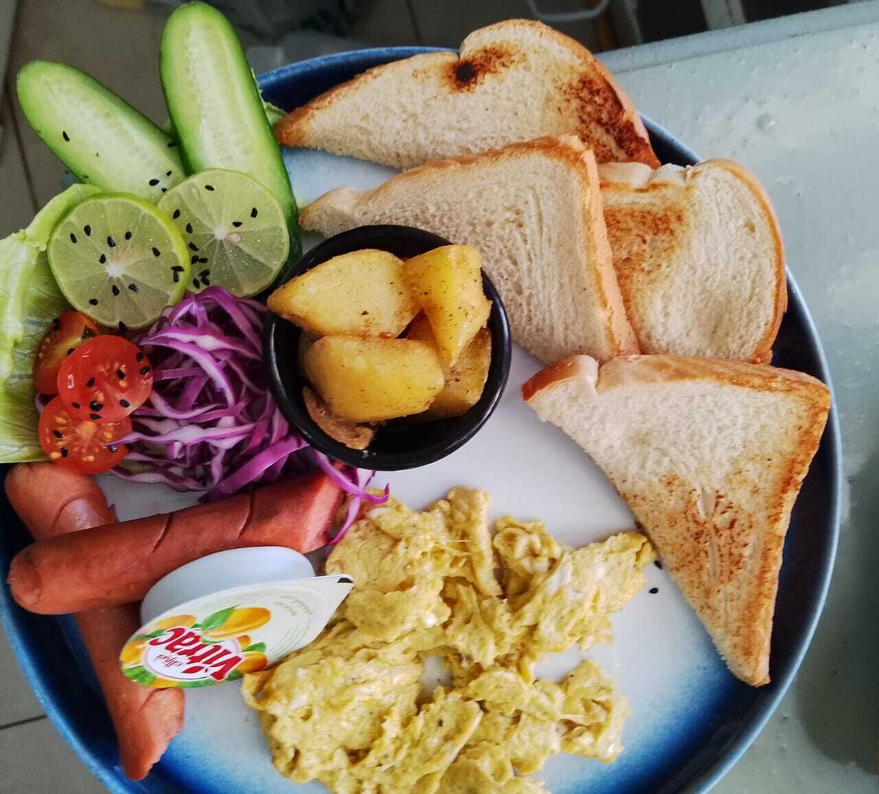 Une assiette contenant des œufs brouillés, des toasts, des tranches de concombre, des oignons rouges, des tomates cerises et un petit bol de pommes de terre.
