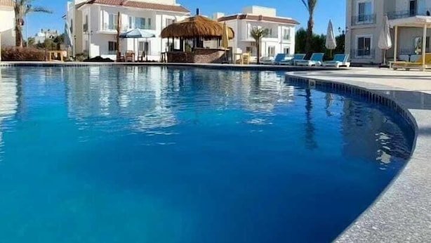 A swimming pool with lounge chairs and a thatched-roof structure, surrounded by white buildings with balconies. - Blue Residence Dahab (Dahab)