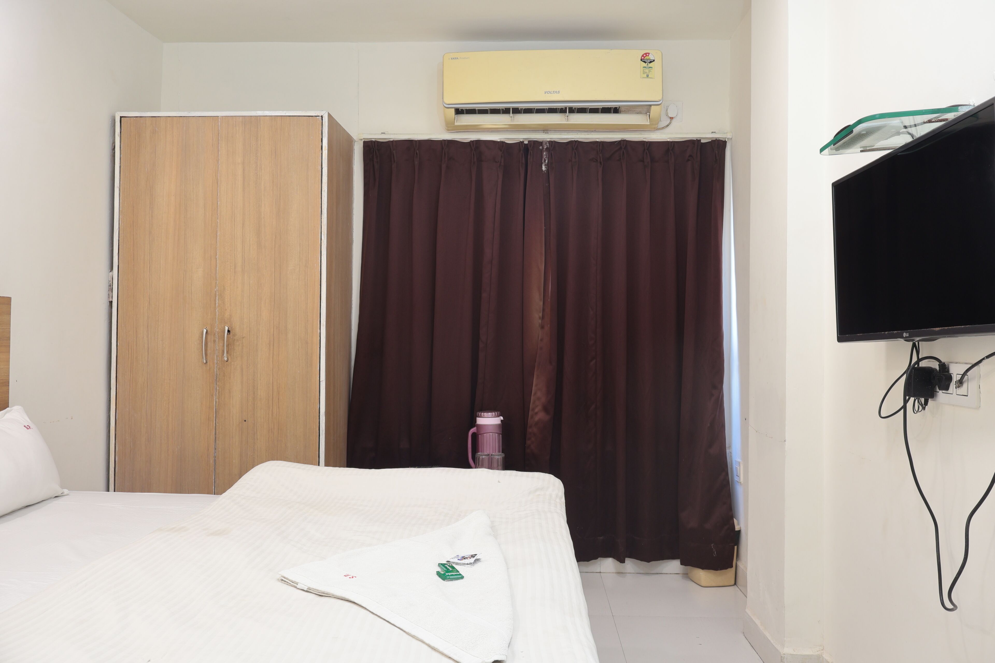 A bedroom with a bed, a wardrobe, an air conditioner, a curtain, a TV mounted on the wall, and a water bottle on the floor.