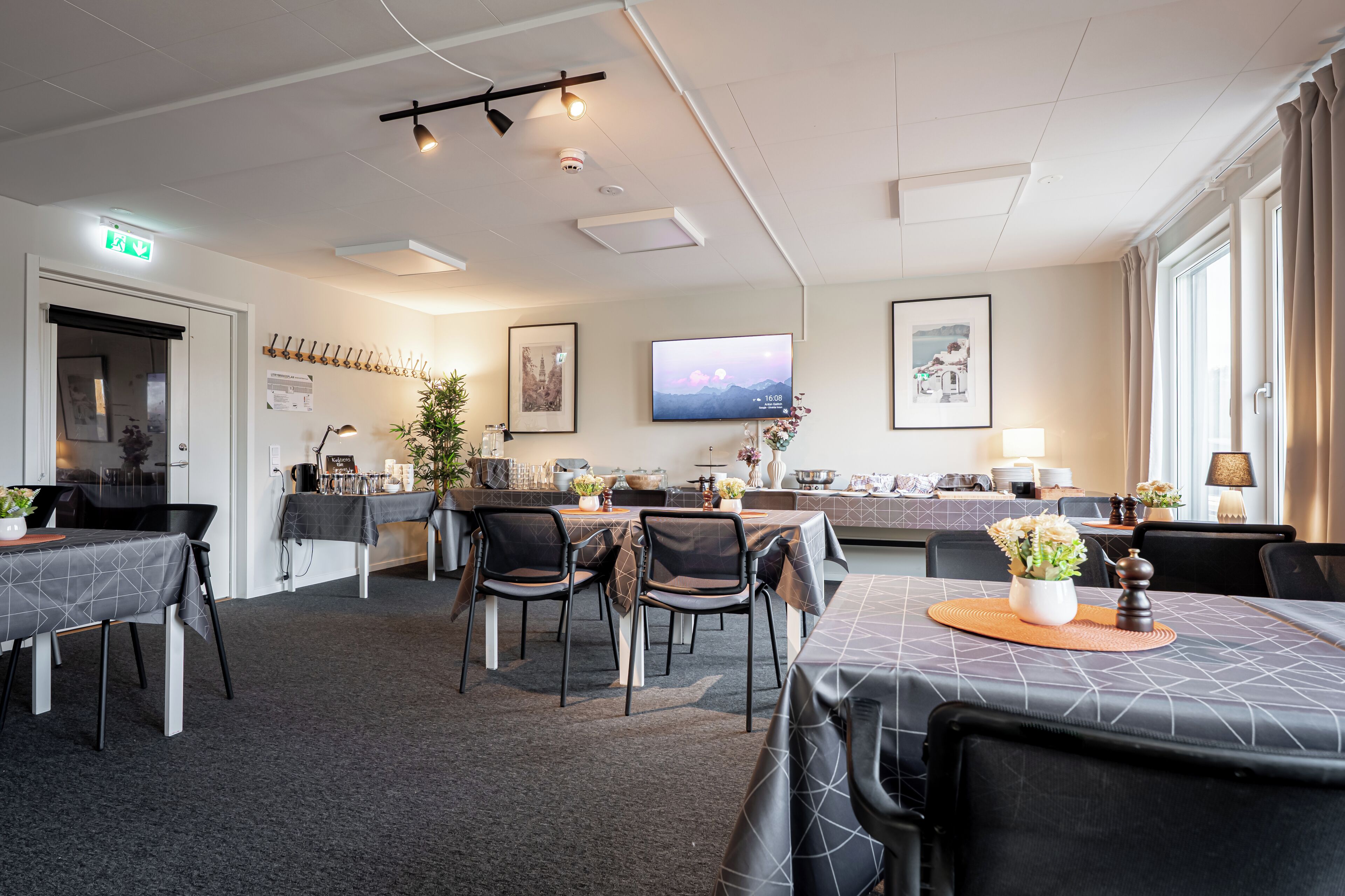 A hotel dining area with tables, chairs, a TV, and a kitchen in the background.