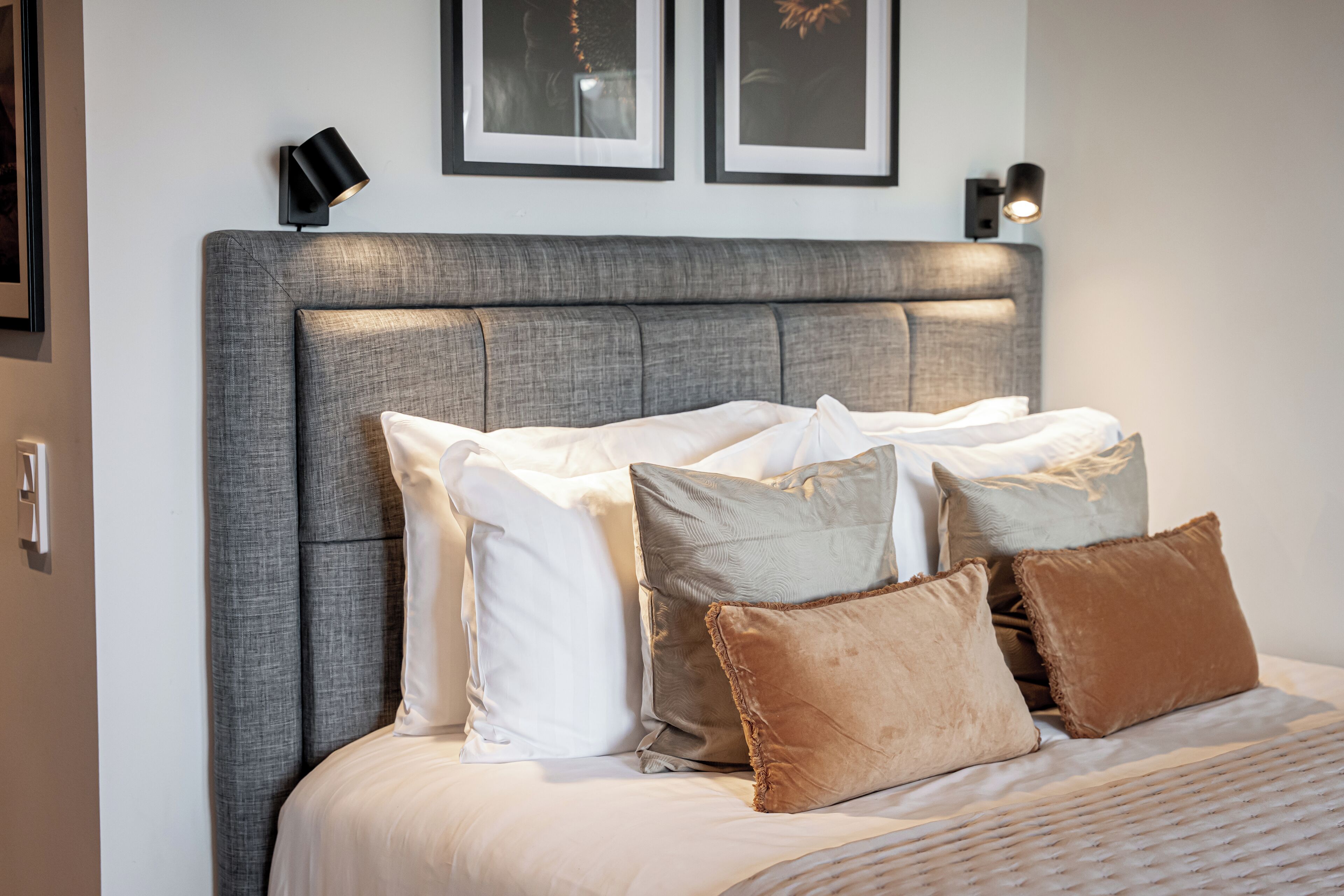 A neatly made bed with a grey upholstered headboard, white and brown pillows, and two wall-mounted reading lamps.