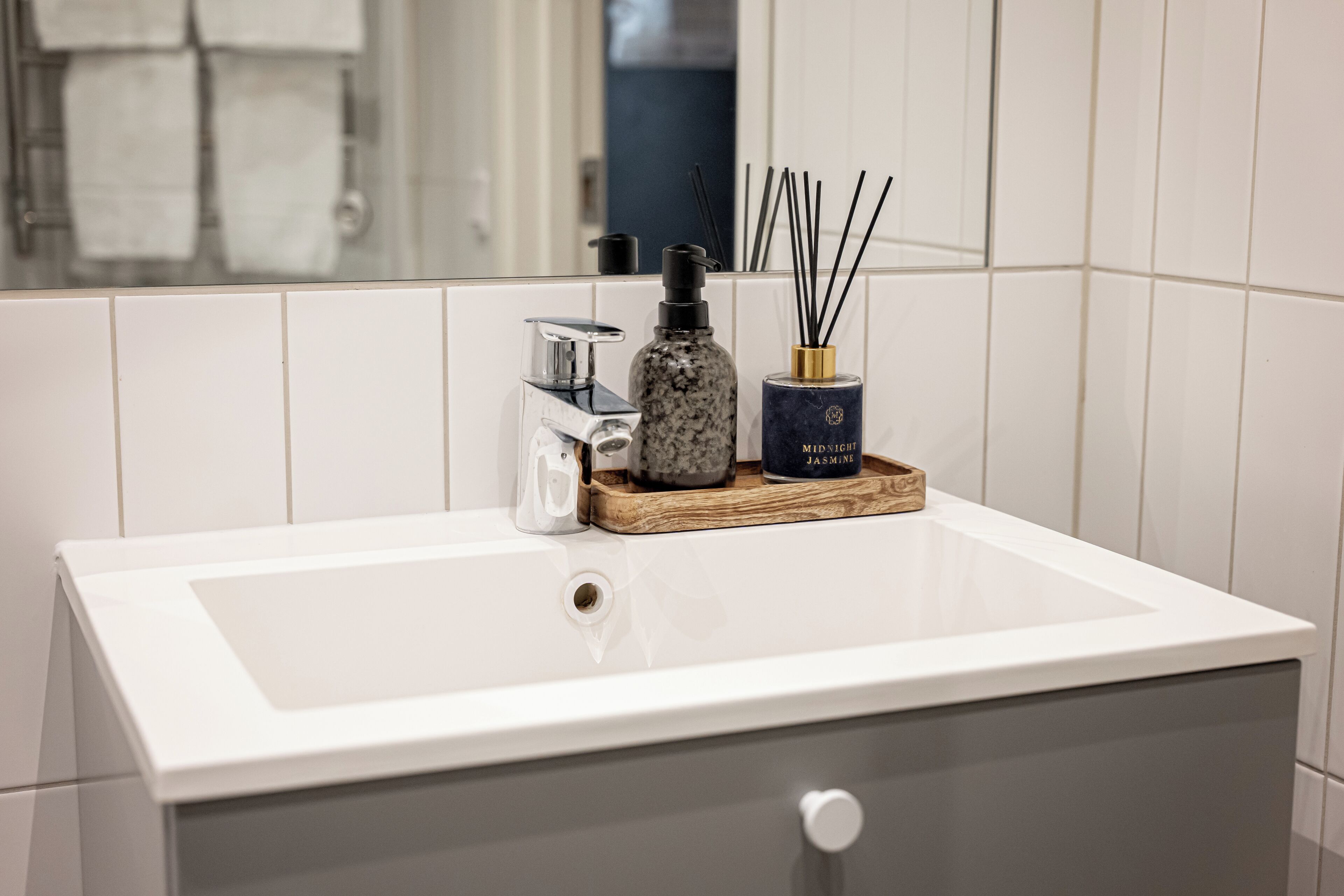 A bathroom with a white sink, a marble soap dispenser, a bottle of reed diffuser, and a wooden tray.