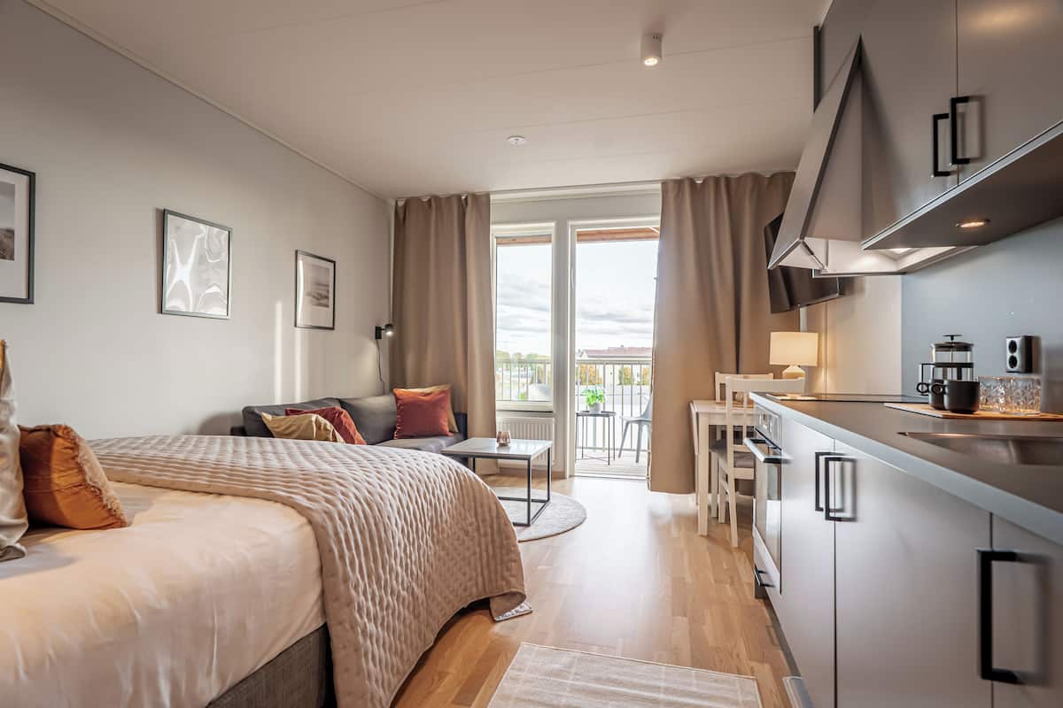 A modern hotel room with a bed, a small table, a kitchenette, and a balcony with a view.