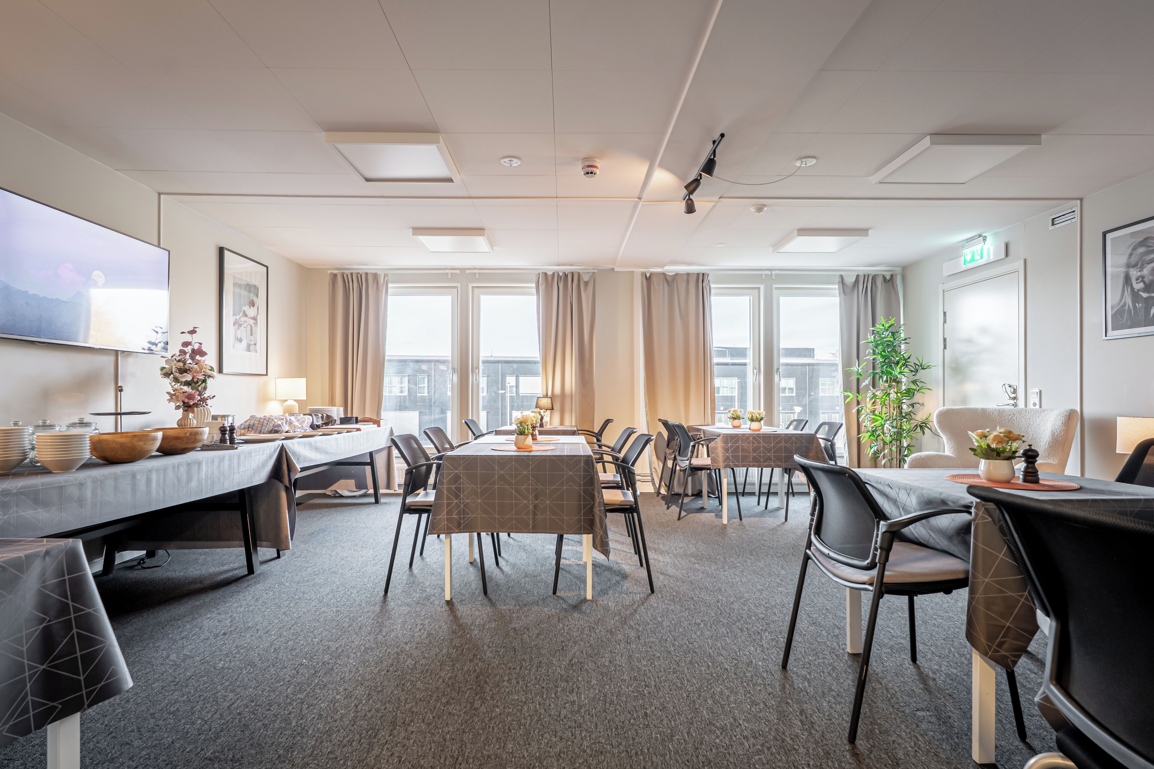 A hotel conference room with multiple tables, chairs, a large flat-screen TV, and a large window with curtains.