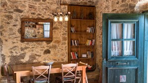 A rustic room with a stone wall, a wooden table with chairs, a bookshelf, and a green door. - Orgon Farm Houses (Apostoli)