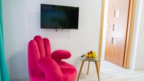 Un soggiorno luminoso con una grande TV a schermo piatto montata a parete, una comoda poltrona a sacco rosa acceso, un tavolino in vetro con della frutta e una porta in legno.