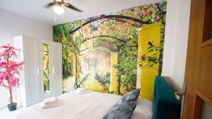 Un dormitorio con un gran mural floral en la pared, una cama con ropa blanca y almohadas estampadas, un ventilador de techo y una planta en maceta.
