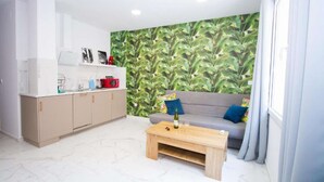 A compact living space with a kitchenette, a grey sofa with colorful cushions, a wooden coffee table, and a green leaf-patterned wall. - Art Apartment IV, Two-Bedroom Apartment (Alicante (Alacant))